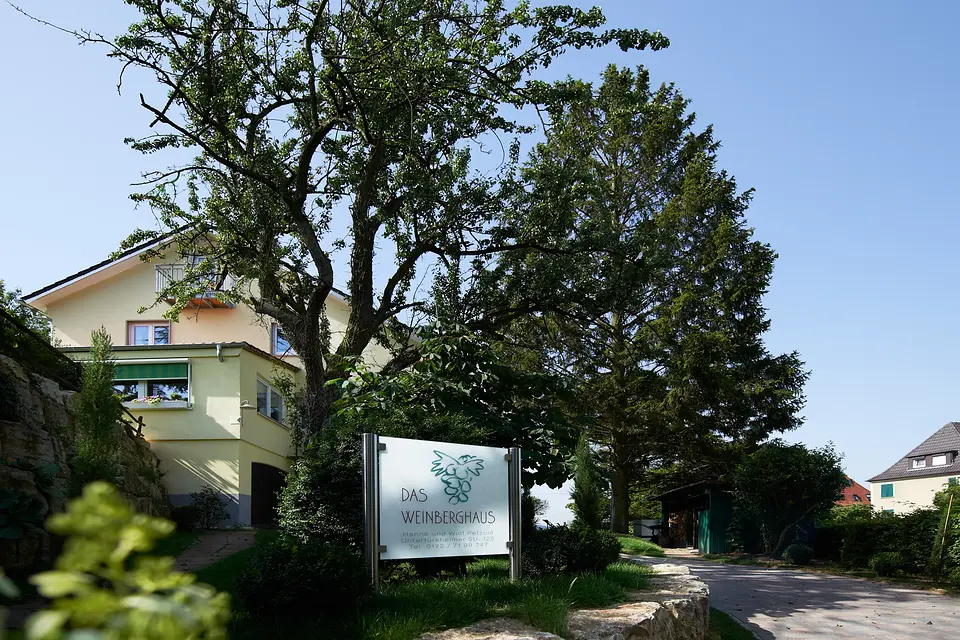 Das Weinberghaus in Fellbach: Namensschild vor dem Haus, umgeben von alten Bäumen und Hecken unter blauem Himmel