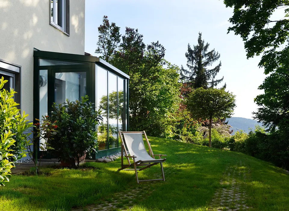 Liegestuhl auf dem Rasen vor dem Wintergarten des Weinberghauses, dahinter Bäume und Blick ins Tal