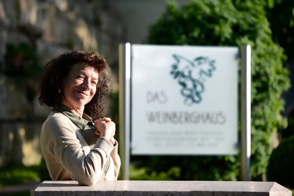 Hanne Petzold lächelt vor dem Schild „Das Weinberghaus“ mit dem Weintrauben-Signet