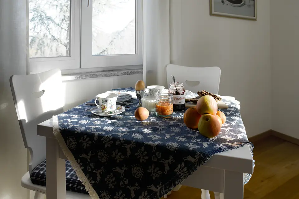 Frühstückstisch am Fenster mit blauer Tischdecke, gekochten Eiern, Äpfeln, Marmelade und Kaffeetasse