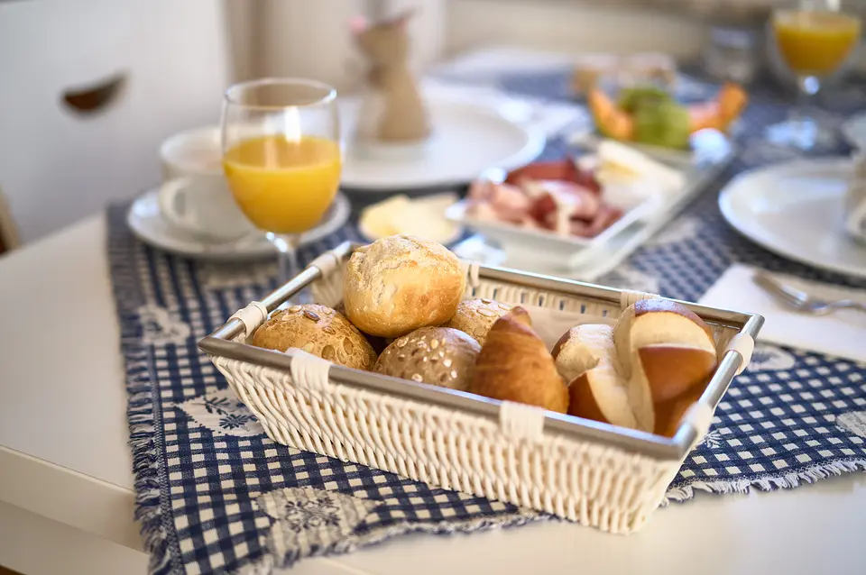 Brötchenkorb mit frischen Brötchen und Croissant auf dem Frühstückstisch, dahinter Orangensaft und Aufschnitt