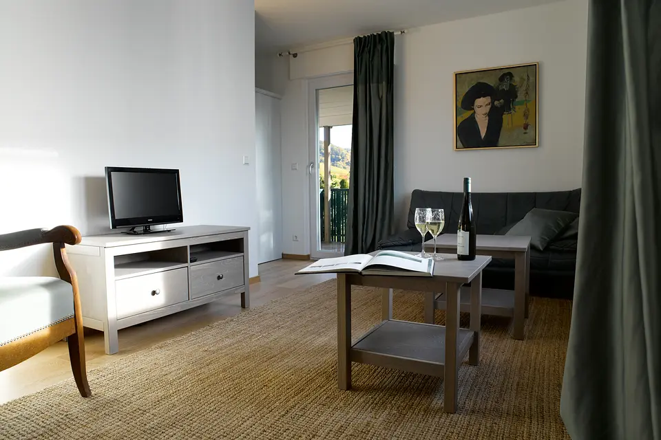 Grünes Zimmer: Sitzecke mit grünem Sofa, Couchtisch mit Weinglas, Fernseher und Balkontür mit Blick ins Grüne