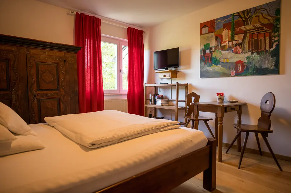 Bauernzimmer: Doppelbett, antiker Bauernschrank, rote Vorhänge, Tisch mit Stühlen, Fernseher und farbiges Ölbild
