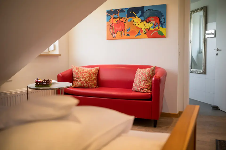 Rote Ledercouch im Weinberg-Zimmer, darüber das bunte Bild mit den springenden Kühen